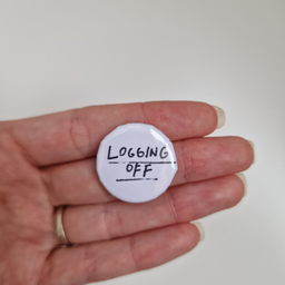 Bedž belog dizajna sa crnim natpisom Logging Off, prikazan u ruci.