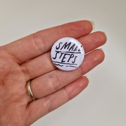 Bedž belog dizajna sa crnim natpisom Small Steps, prikazan u ruci.