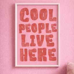 A4 poster sa belim ramom i natpisom Cool People Live Here u roze i crvenim tonovima na pastelno roze zidu.