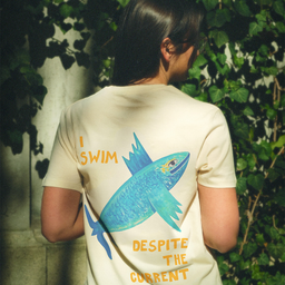 Devojka nosi pamučnu majicu na kratke rukave, bež boje, sa ilustracijom ribe i natpisom: I SWIM DESPITE THE CURRENT