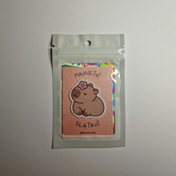 Magnet sa ilustracijom kapibare sa ružičastim cvetićem na glavi, spakovan u providnu kesicu sa holografskim ivicama.