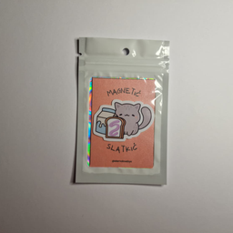 Magnet sa ilustracijom sive mace koja sedi pored parčeta torte u kutiji, spakovan u providnu kesicu sa holografskim ivic