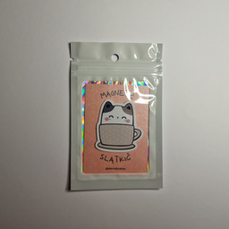 Magnet u obliku bele mace sa crnim ušima koja sedi u bež šoljici sa tufnicama, spakovan u providnu kesicu sa holografski