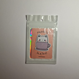 Magnet sa ilustracijom bele mace koja sedi u ljubičastoj šoljici sa tufnicama, spakovan u providnu kesicu sa holografski