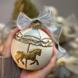 Dekorativna sveća u obliku novogodišnje kugle sa zlatnim konjićem i mašnom, držana u rukama, sa zamućenom prazničnom poz