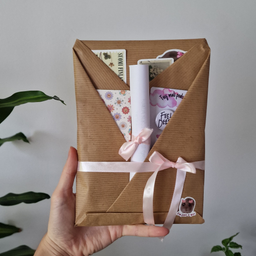 Ruka drži set roze dnevnog planera i To Do liste, okružene biljkama u pozadini. Prikazan je poklon paket koji sadrži pla